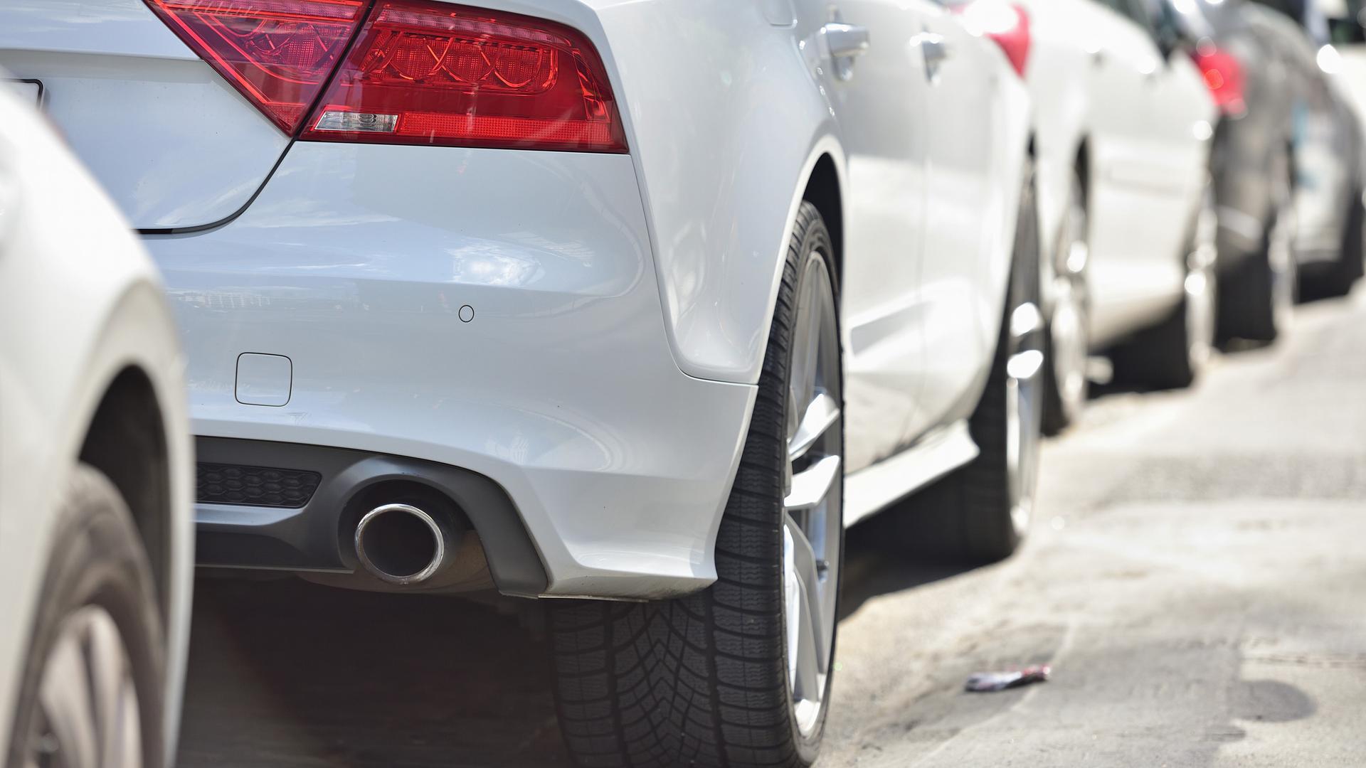 Mehrer Autos stehen aufgereiht am Straßenrand, beim ersten in der Reihe ist am Heck ein Auspuff zu sehen.