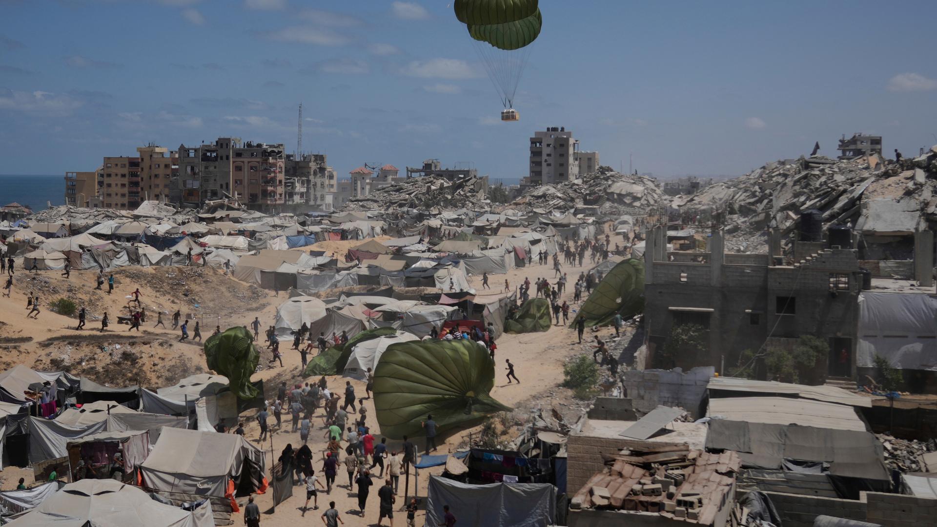 Palästinenser sammeln Hilfsgüter ein, die am 7. August 2025 mit Fallschirmen über Gaza-Stadt im nördlichen Gazastreifen abgeworfen werden. Im Hintergrund Fallschirme, Zelte, zerstörte Gebäude und das Mittelmeer.