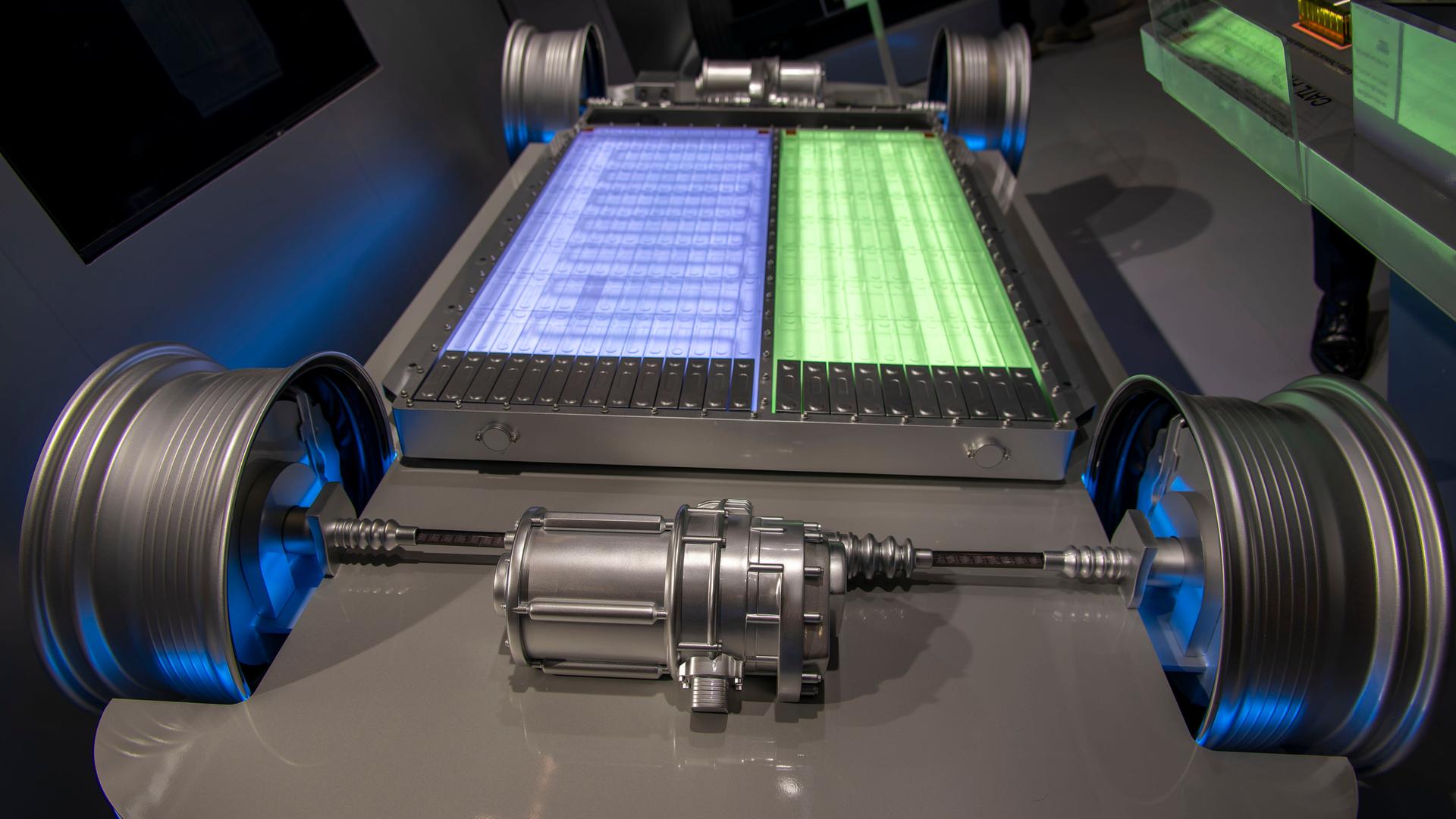 Zu sehen ist der Unterboden eines Elektrofahrzeugs ohne Karosserie, so dass die Batterie frei sichtbar ist. In der Mitte liegt ein großes, rechteckiges Batteriepaket, das aus zwei unterschiedlich farbigen Bereichen besteht.