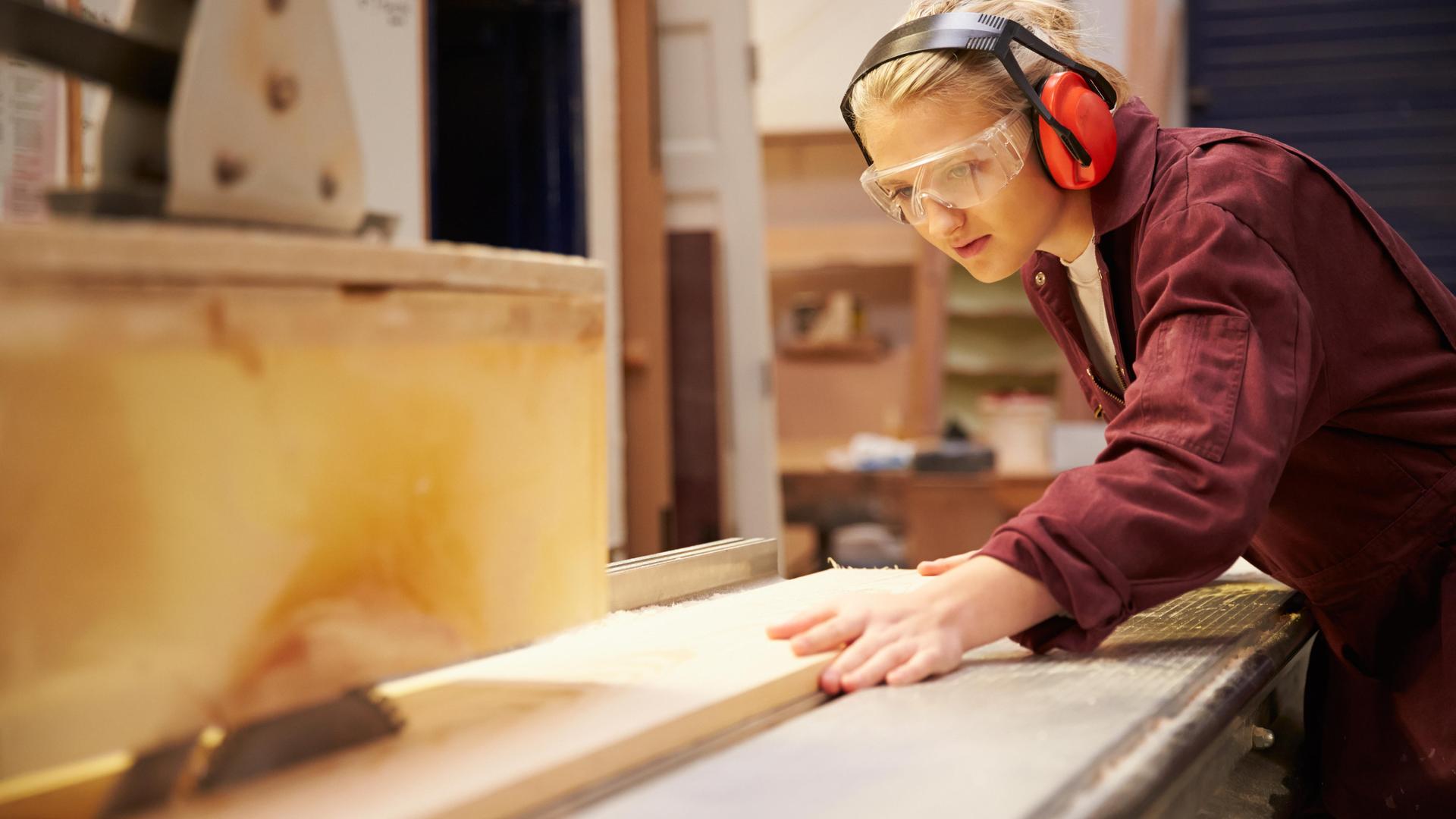 Eine junge Frau in Arbeitskleidung und mit Schutzbrille arbeitet an einer Kreissäge 