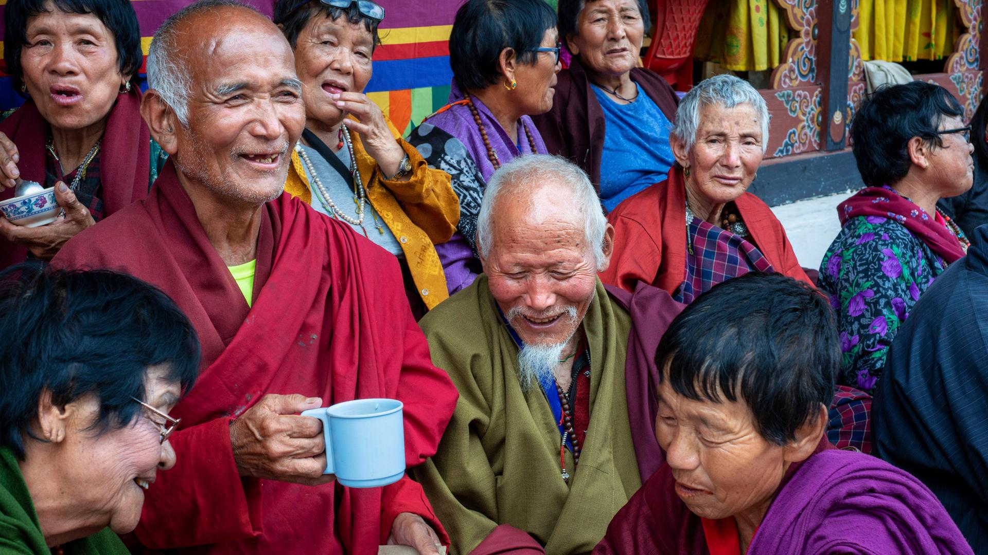 Gläubige Buddhisten in traditioneller Kleidung. Wollene Mäntel in Dunkelrot und Olivgrün. Die älteren Menschen unterhalten sich, viele lächeln. Ein Mann hält eine Tasse in der Hand. Alle machen einen zufriedenen Eindruck
