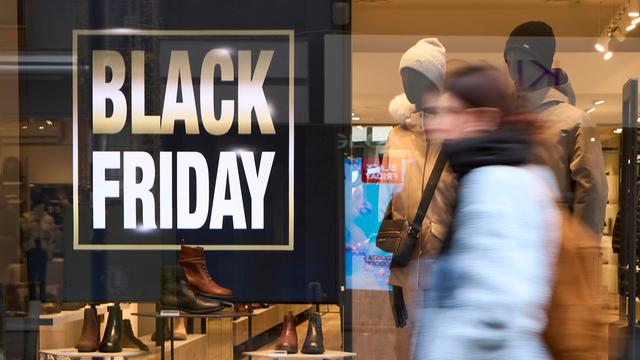 Fußgänger in Winterkleidung laufen an einem hell erleuchteten Schuhladen vorbei; im Schaufenster sind ein großes „BLACK FRIDAY“-Schild und Lederschuhe zu sehen.