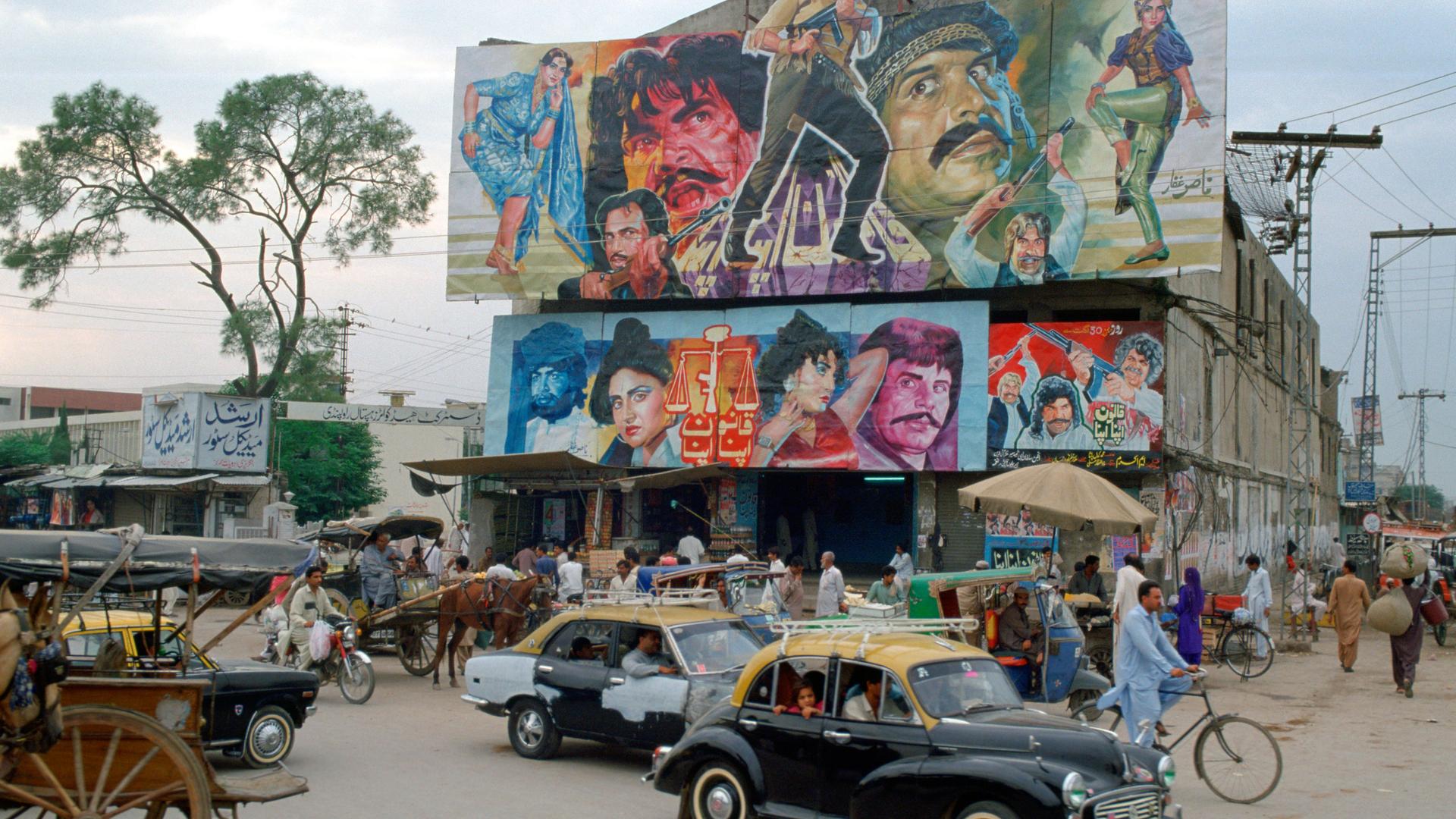 Belebte Straßenecke mit Gebäude, auf dessen Dach ein großes Filmplakat mit bunt gekleideten Figuren angebracht ist, das für einen Bollywoodfilm wirbt.
