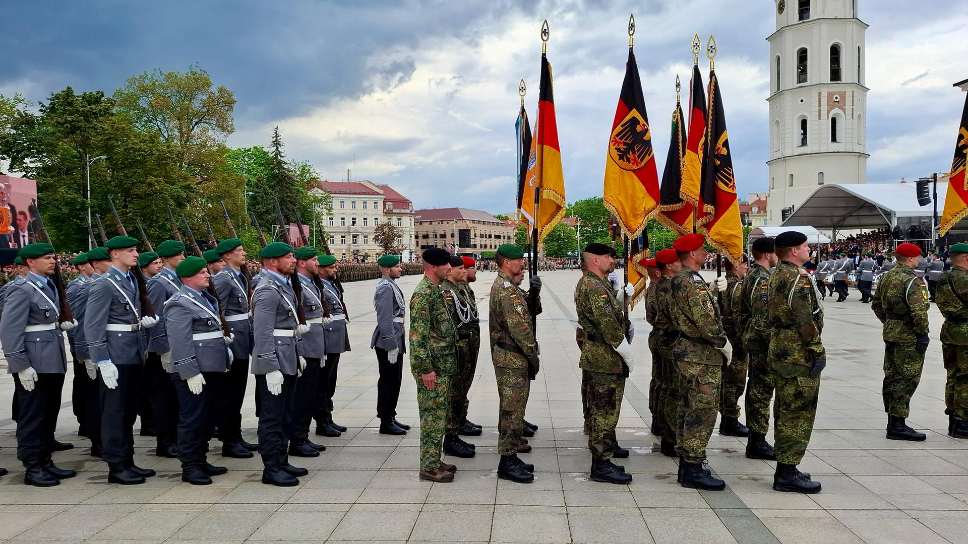 Deutsche Soldaten stehen in Uniformen auf einem öffentlichen Platz in Litauen, einige halten die deutsche Flagge in der Hand. Im Hintergrund ist der Glockenturm der Kathedrale St. Stanislaus in Vilnius zu sehen.