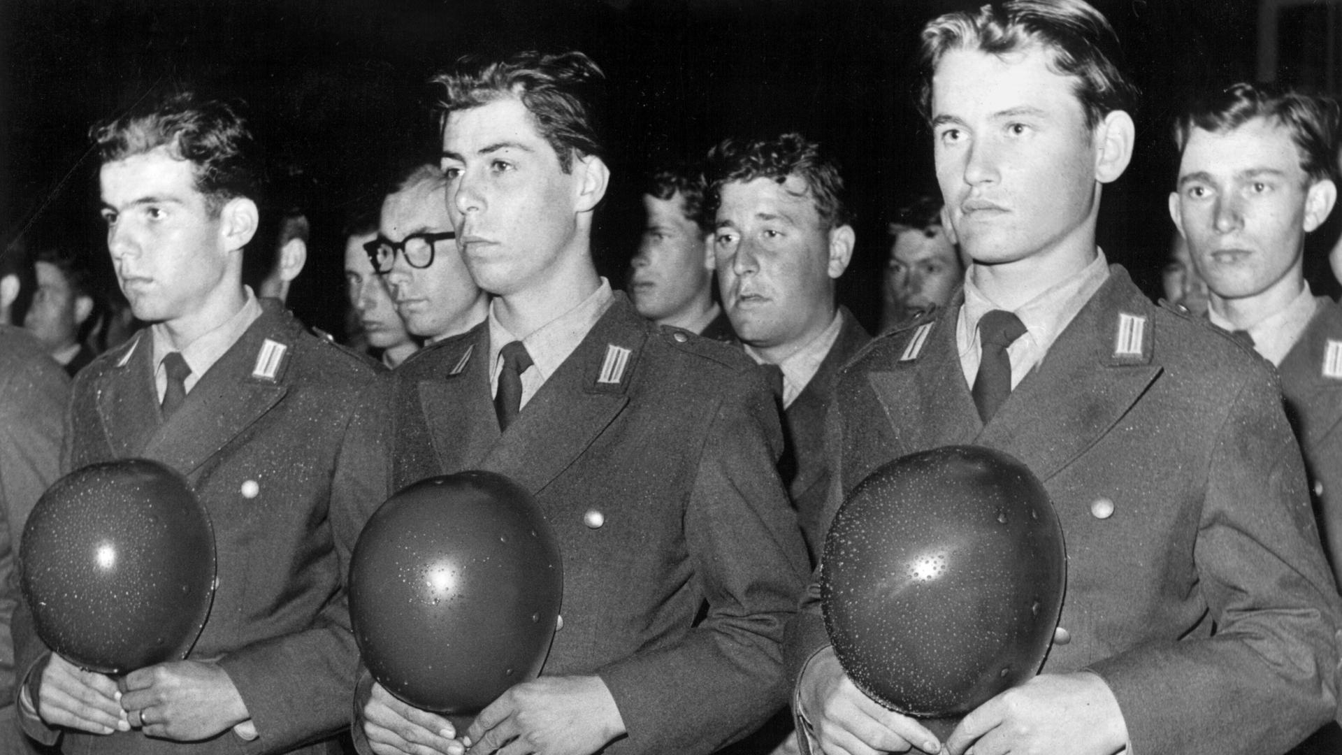 Historisches Schwarzweißfoto von Bundeswehrrekruten während der feierlichen Verpflichtung. Sie stehen aufgereiht nebeneinander und halten ihre Helme vor der Brust.