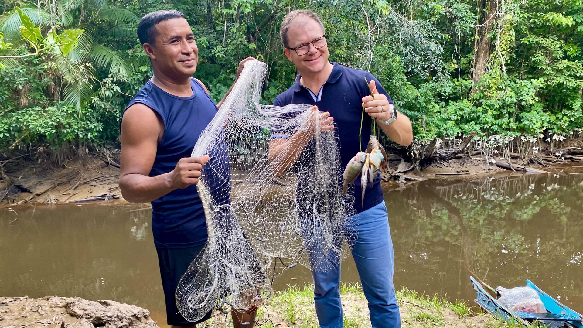 Bundesumweltminister Carsten Schneider (r, SPD) lässt sich von einem Bewohner der Quilombola-Gemeinschaft Abacatal Fische zeigen, die der örtliche Lehrer eben mit einem Netz im Fluss gefangen hat.