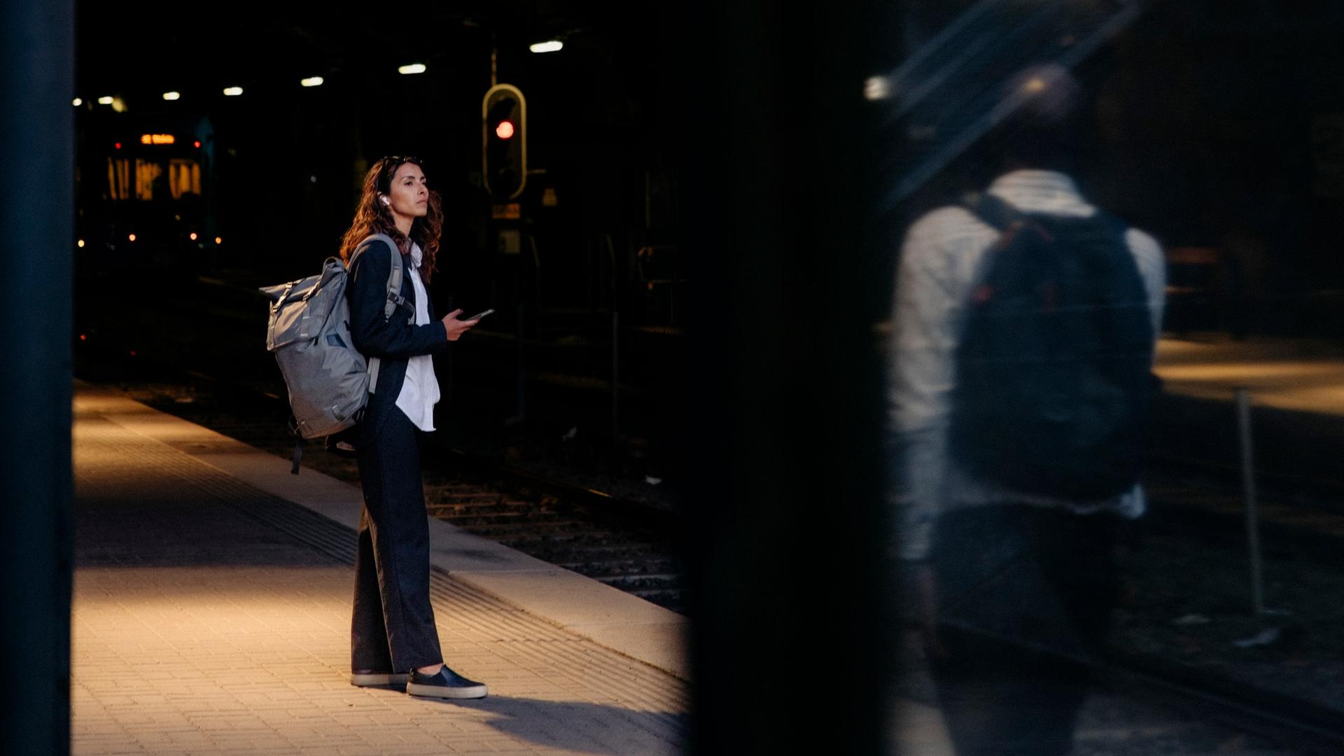 Eine Frau steht an einem Bahnhof. Unscharf im Bildvordergrund steht ein Mann, der sie beobachtet.