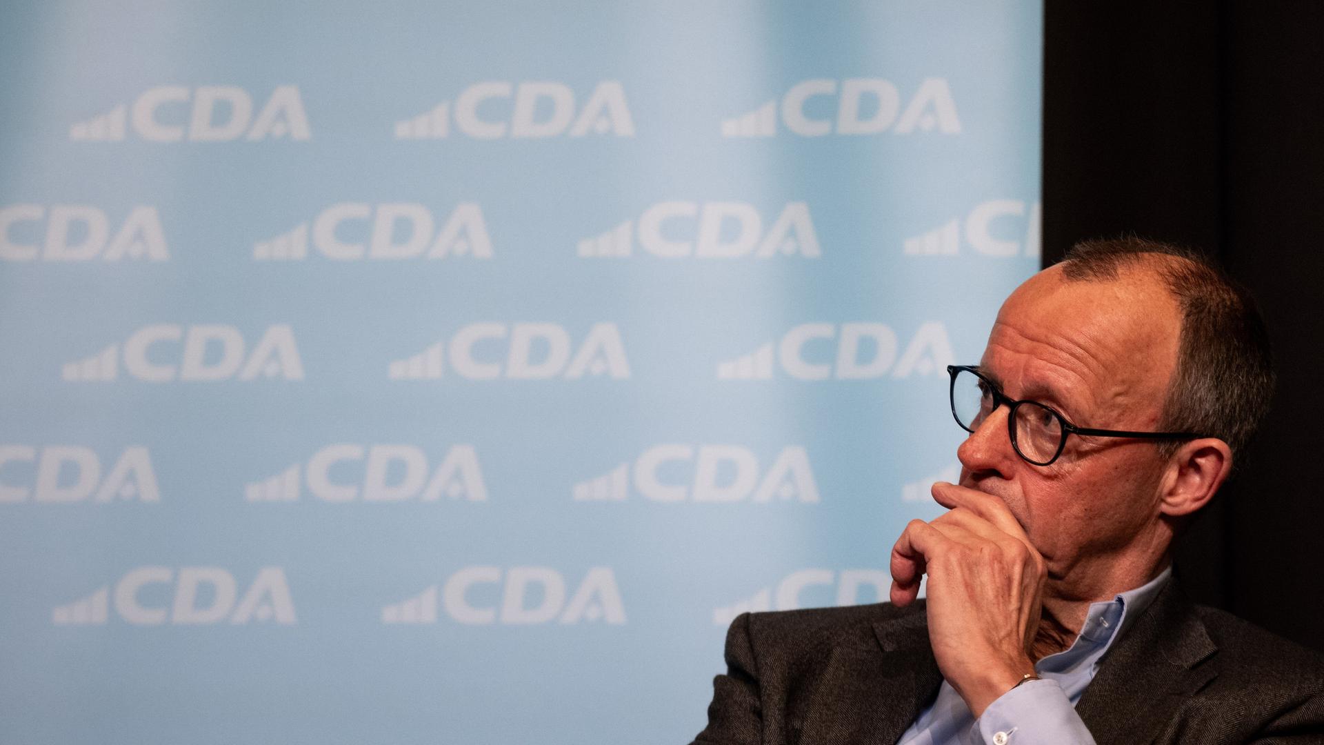 Friedrich Merz sitzt bei einer Veranstaltung mit nachdenklicher Miene vor einer Wand mit dem CDA-Logo.