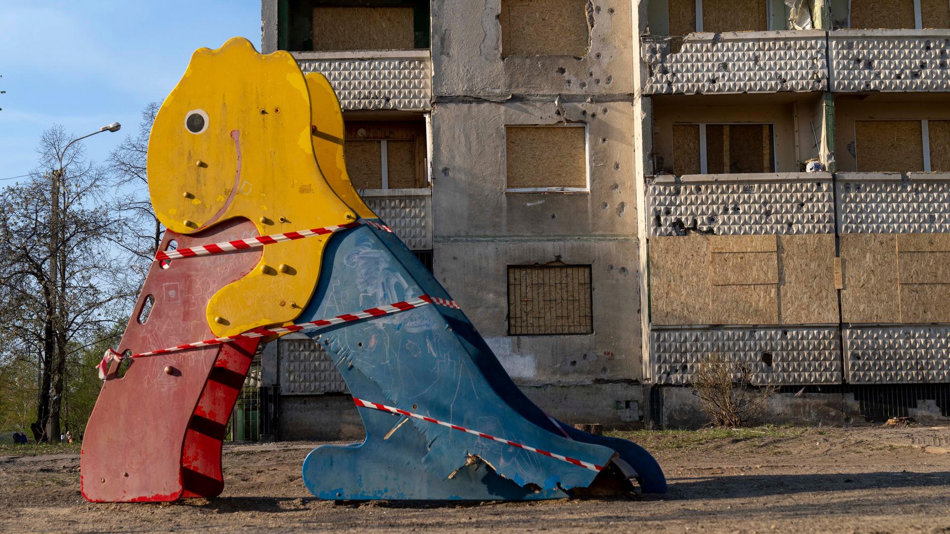 Auf einem Spielplatz steht eine beschädigte bunte Rutsche in Form eines Dinosauriers, im Hintergrund ist ein vom Krieg stark beschädigtes Gebäude zu sehen.