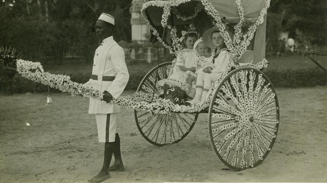 Ein Diener zieht eine mit Blumen geschmückte Rikscha mit drei weißen Kindern am vom Frauenverein des Roten Kreuzes organisierten "Margaritentag" in Daressalam (1912).