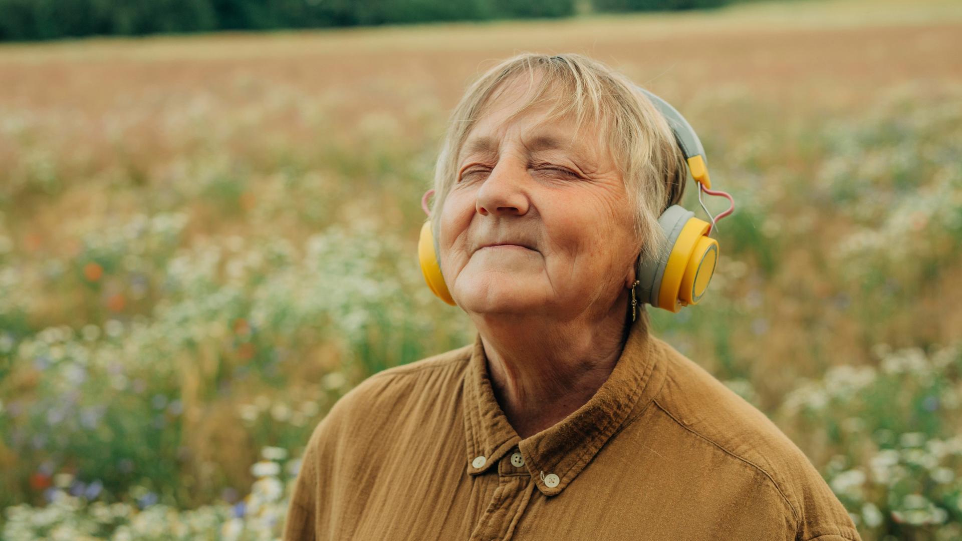 Eine ältere Frau steht inmitten eines Feldes, trägt große gelbe Kopfhörer und lächelt entspannt mit geschlossenen Augen. Sie wirkt versunken in die Musik.