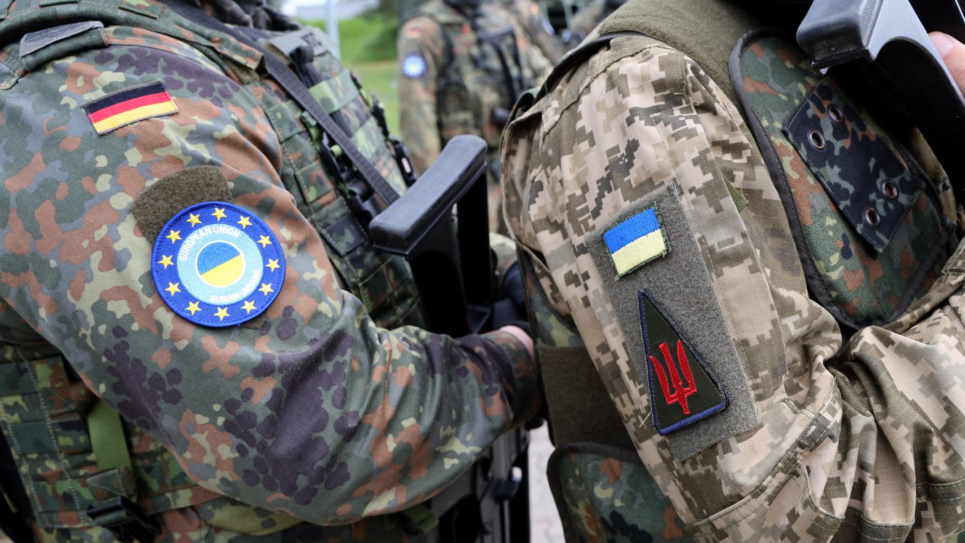 Schulter an Schulter stehen ein deutscher und ein ukrainischer Soldat auf einem Truppenübungsplatz. Man sieht nur die Uniform.