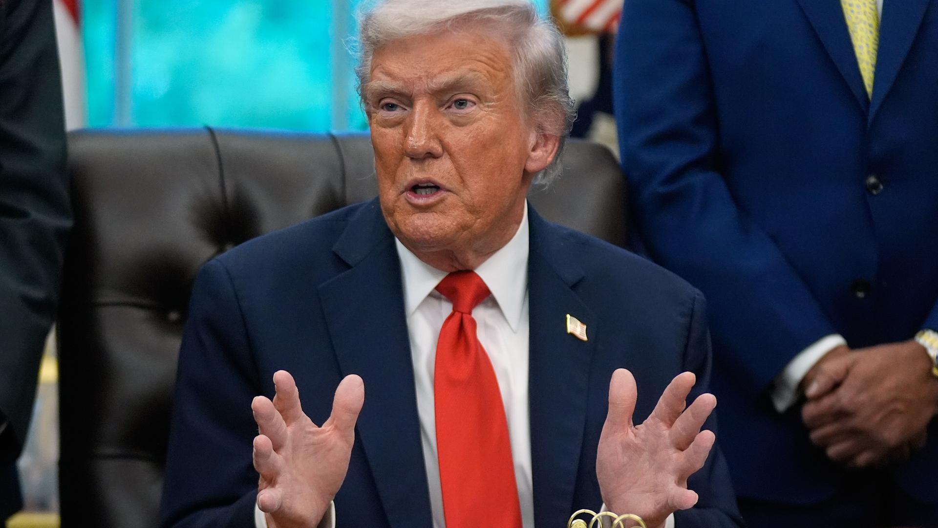 Donald Trump sitzt an seinem Schreibtisch im Oval Office und breitet die Hände aus.