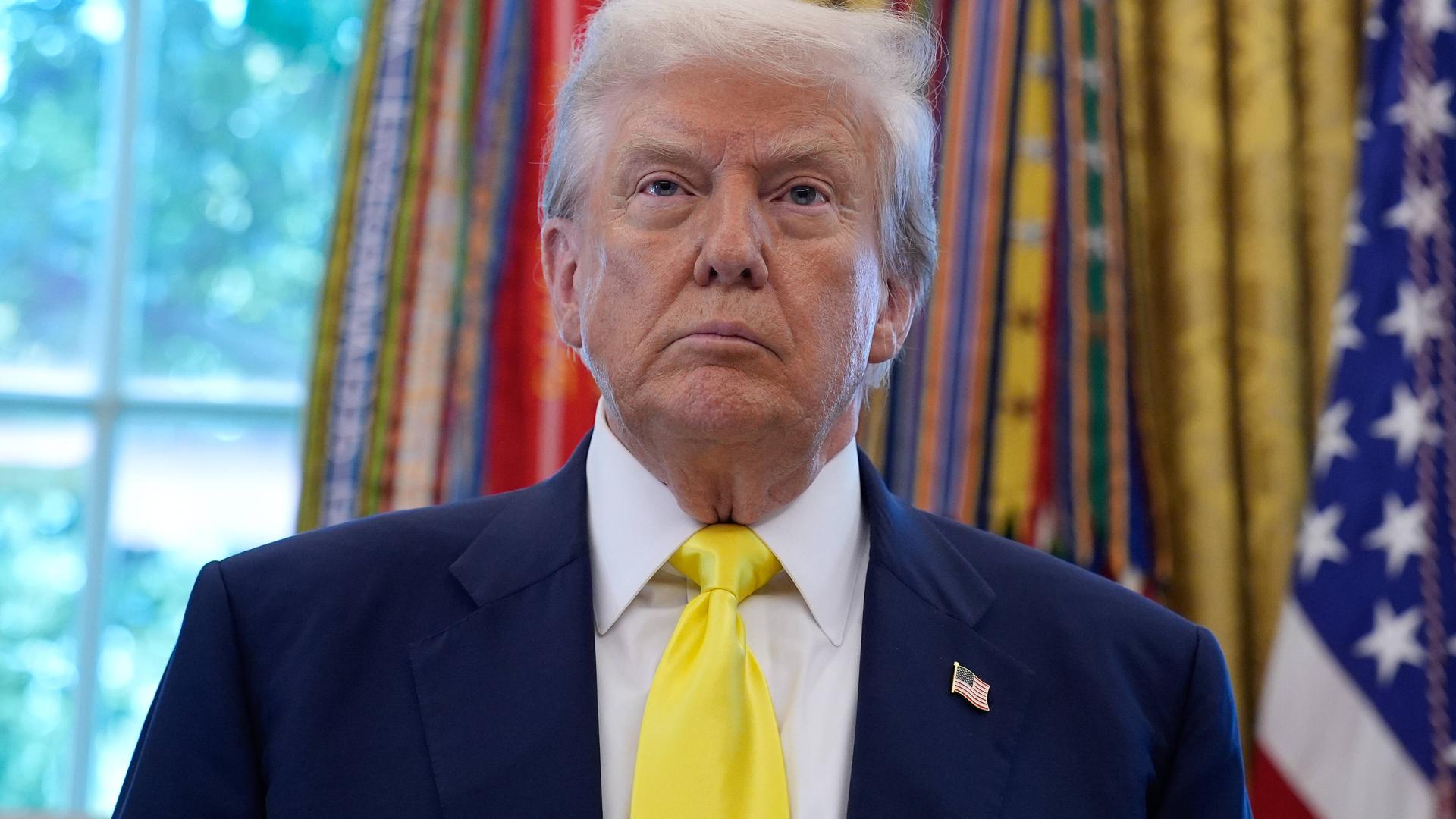 US-Präsident Donald Trump steht vor den Fahnen im Oval Office des Weißen Hauses in Washington und schaut mit ernstem Gesichtsausdruck