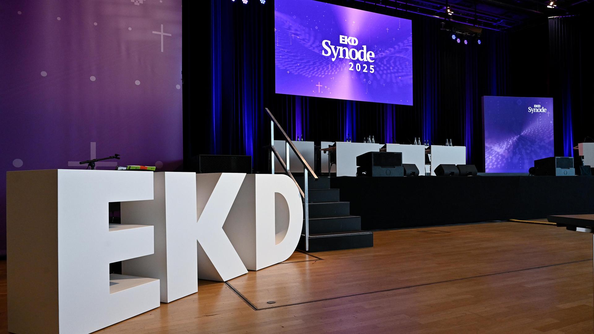 Die drei Buchstaben EKD stehen vor dem leeren Podium, auf dem Bildschirm über dem Podium ist "EKD Synode 2025" zu lesen.