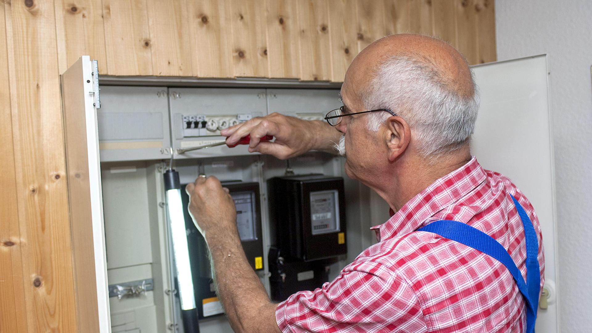 Ein Mann mit Glatze und blauer Latzhose prüft an einem Stromkasten die Leitungen