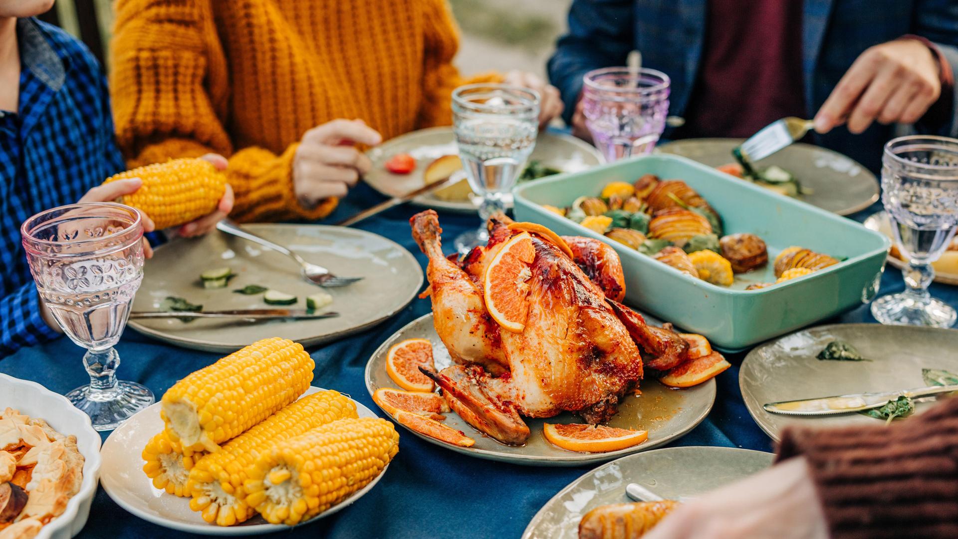 An einem reich gedecktem Tisch mit Maiskolben, Geflügelbraten und Geschirr sitzen mehrere Personen und essn