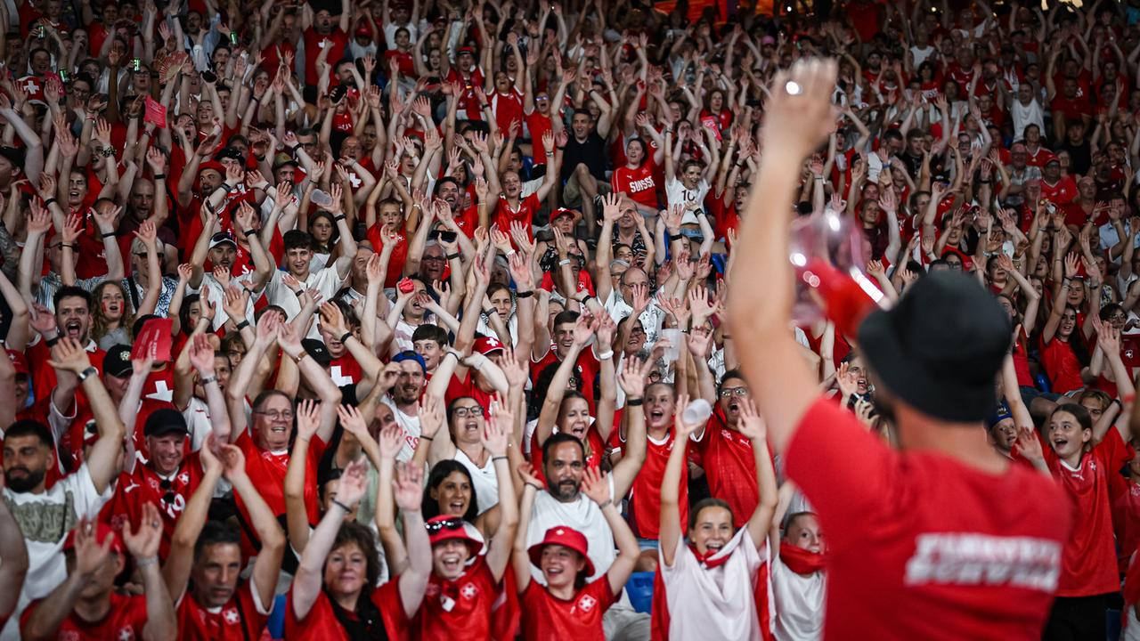 Schweizer Fans beim Eröffnungsspiel gegen Norwegen. Die Zuschauerinnen und Zuschauer heben fröhlich die Hände zu La Ola, sie tragen Schweizer Trikots. 