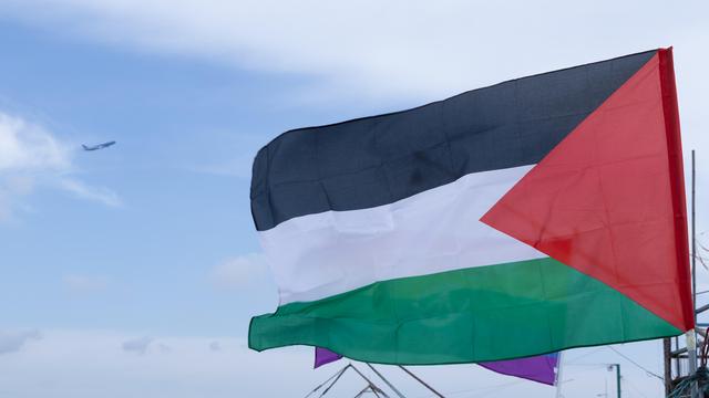 Die Palästinensische Flagge weht an einem Mast. Sie ist horizontal in den panarabischen Farben Schwarz, Weiß und Grün unterteilt. An der linken Seite ist ein rotes Dreieck.