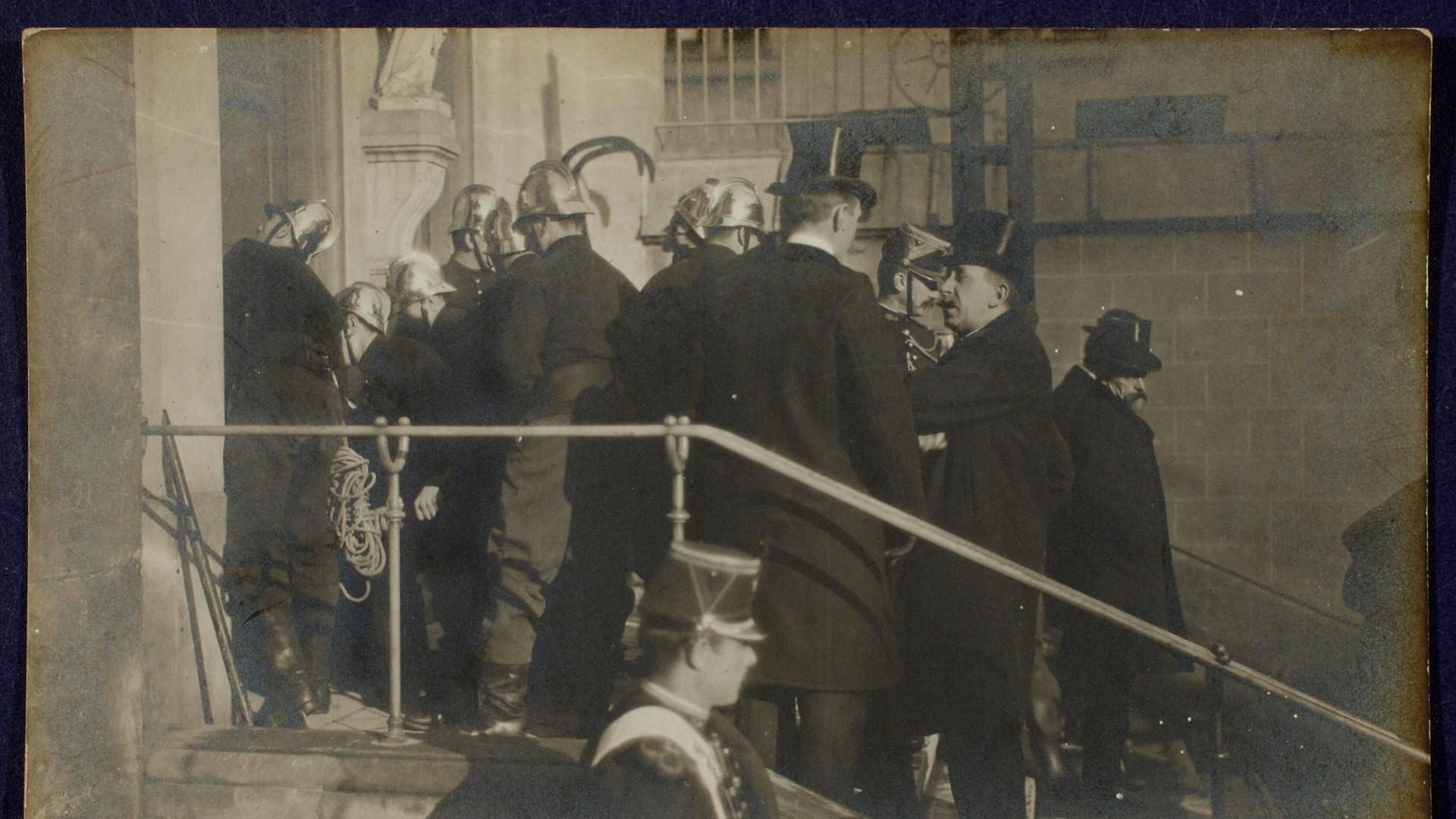 Szene auf einer Fotopostkarte: Polizisten und Feuerwehrleute in Uniformen und Helmen stehen auf einer Treppe vor einer Kirche. Sie sind dabei, die Türen aufzubrechen.