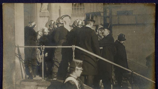 Szene auf einer Fotopostkarte: Polizisten und Feuerwehrleute in Uniformen und Helmen stehen auf einer Treppe vor einer Kirche. Sie sind dabei, die Türen aufzubrechen.