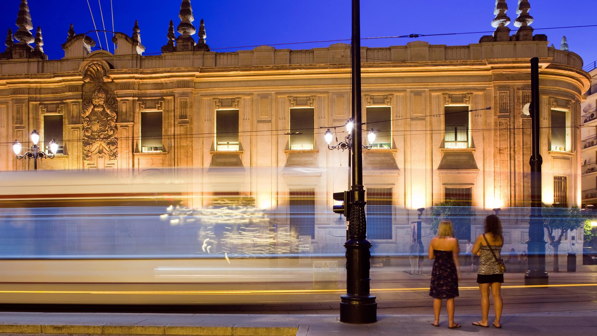 Zwei junge Frauen stehen am Abend in Sevilla vor einer vorbeifahrenden Straßenbahn, im Hintergrund ist ein erleuchtetes Gebäude zu sehen.