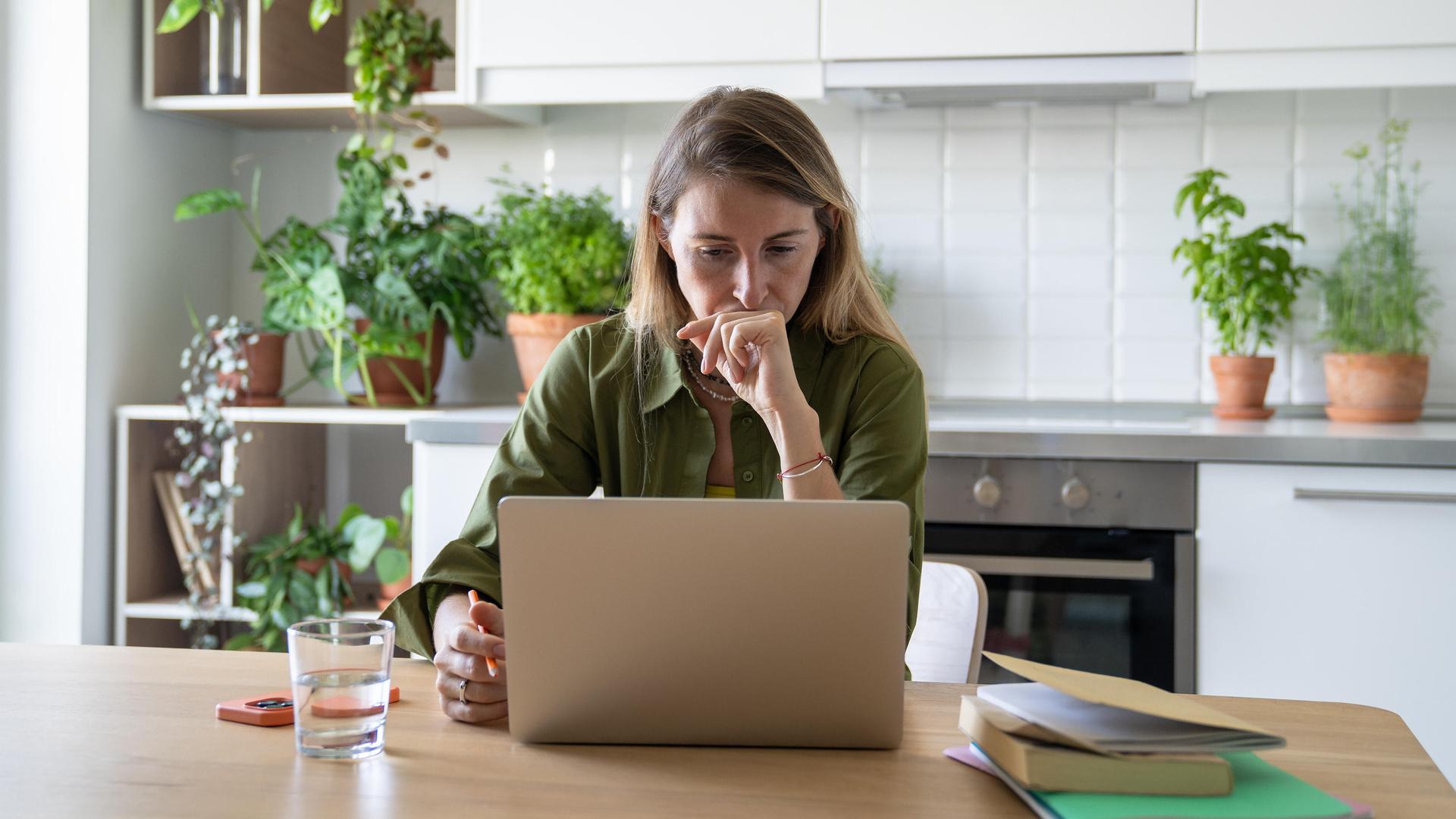 Eine Frau sitzt am Küchentisch an ihrem Laptop; neben ihr ein Handy, ein Wasserglas und ein Stapel Hefte und Bücher. 