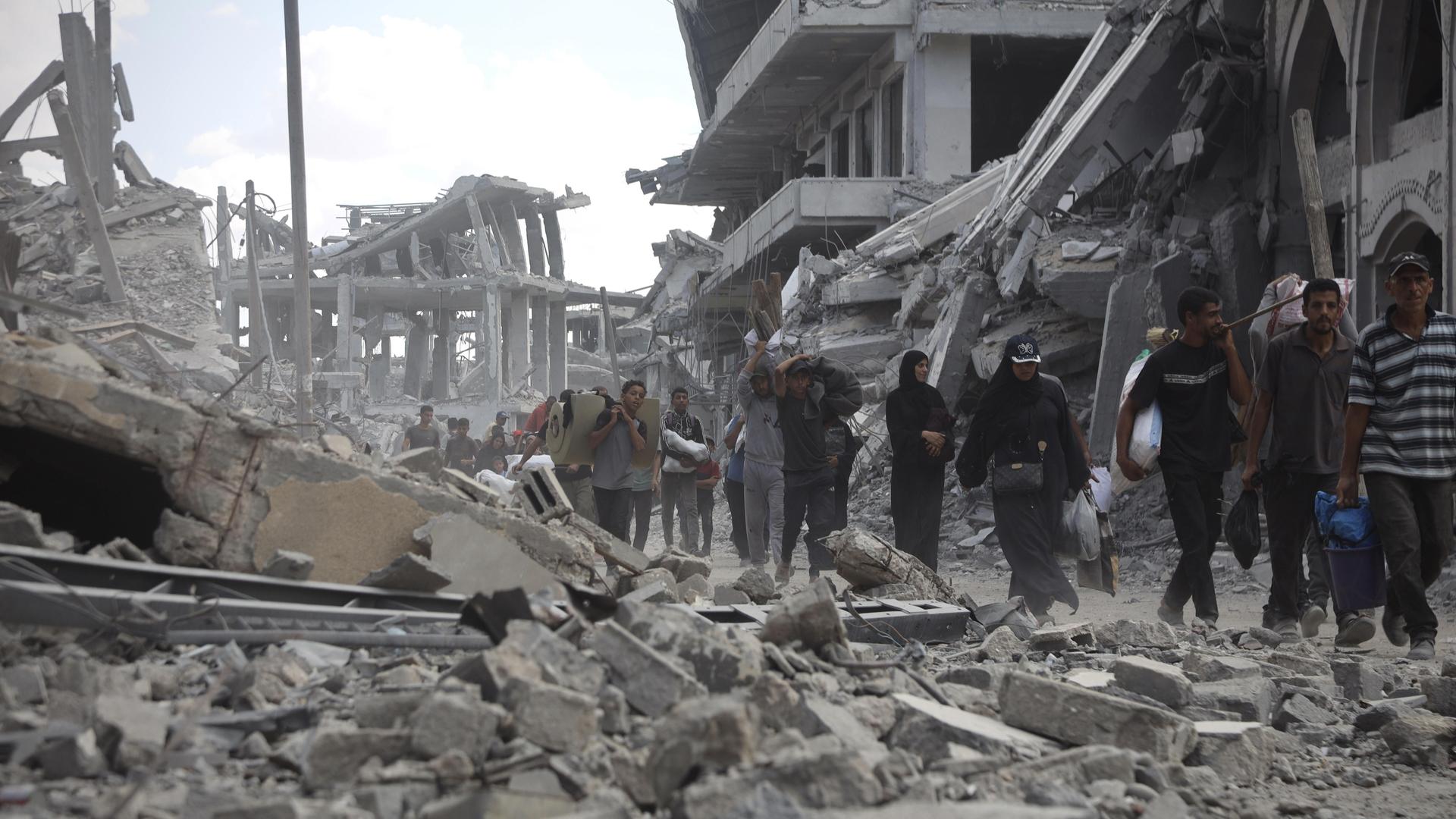 Mehrere Menschen laufen durch eine Straße voller Schutt und eingestürzter Gebäude. Im Hintergrund sind schwer beschädigte Betonbauten zu sehen. 