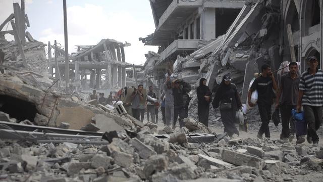 Mehrere Menschen laufen durch eine Straße voller Schutt und eingestürzter Gebäude. Im Hintergrund sind schwer beschädigte Betonbauten zu sehen. 