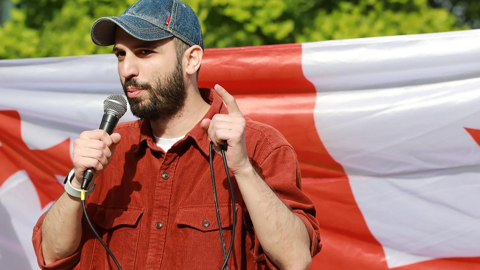 Giorgi Kakabadze steht mit einem Mikrofon in der Hand vor einer georgischen Flagge und spricht