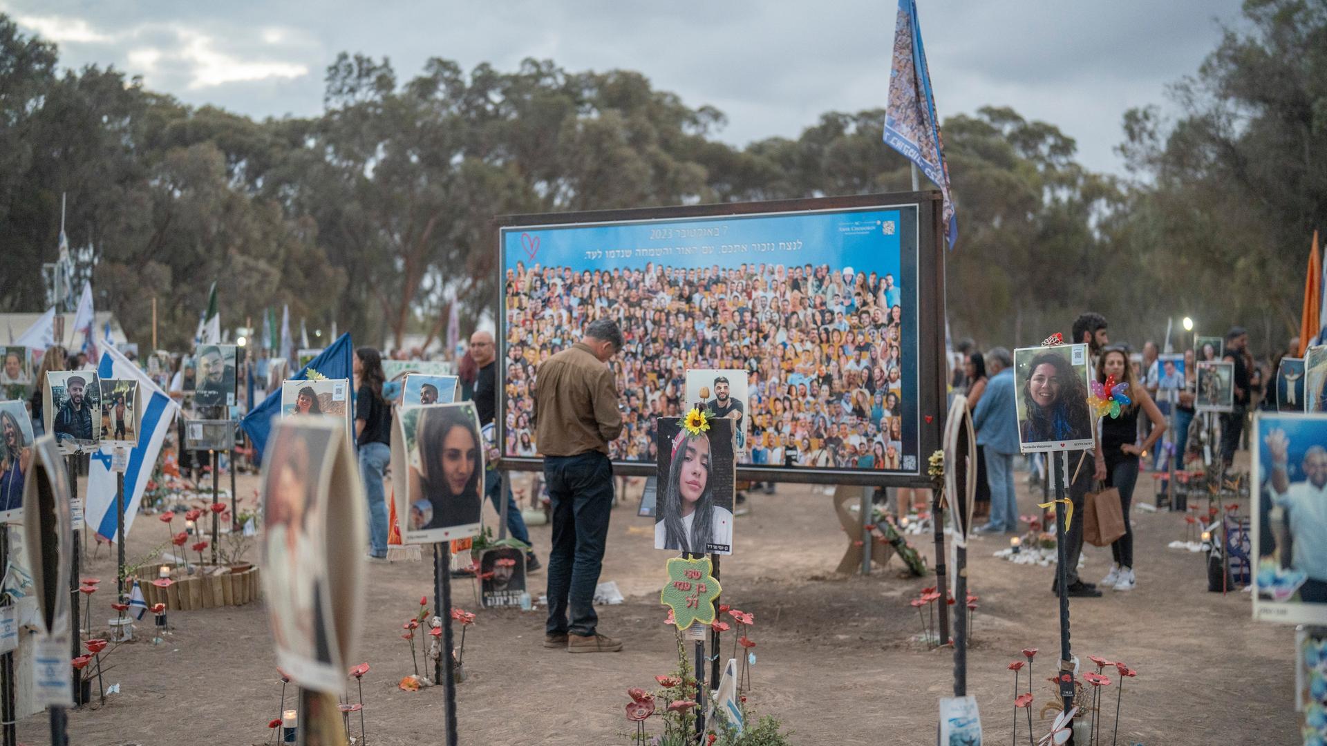 Porträtfotos von Opfern des Hamas-Angriffs sind an Stangen befestigt, die in der Erde stecken. Vor einer Tafel mit Bildern der Getöteten steht ein trauernder Mann.