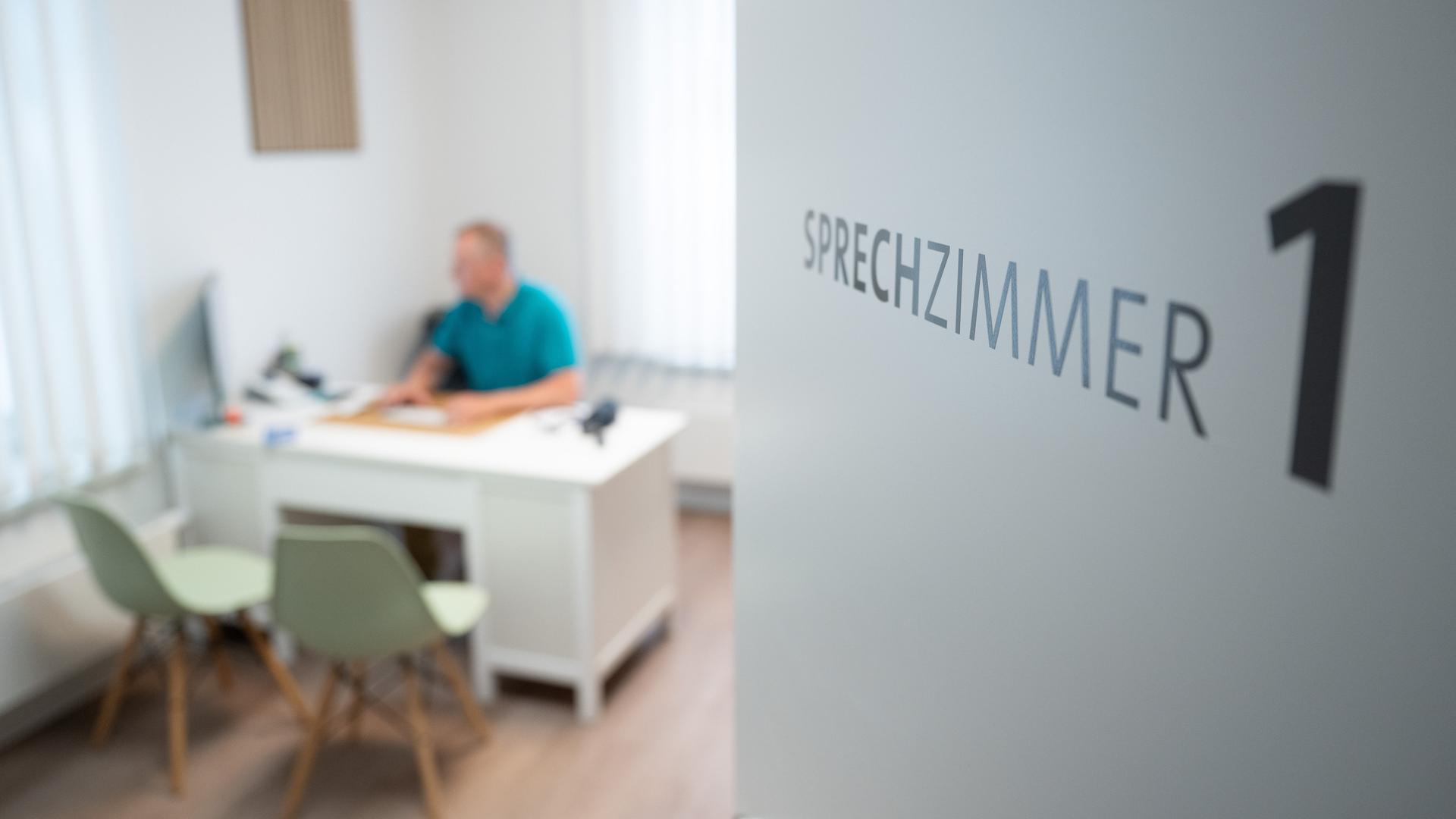 Ein Hausarzt sitzt in einem Sprechzimmer seiner Hausarztpraxis an einem Schreibtisch und arbeitet am Computer. 