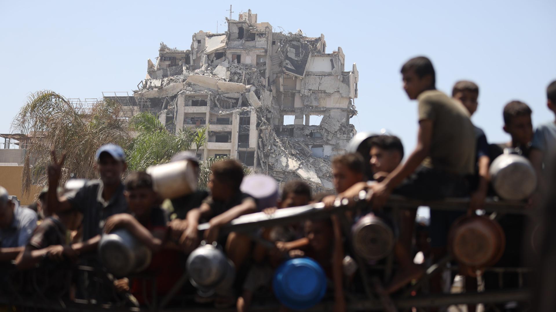 Palästinenser, darunter auch Kinder versammeln sich in Gaza-Stadt, um warme Mahlzeiten entgegenzunehmen, die von der Wohltätigkeitsorganisation verteilt werden. Im Hintergrund ist ein zerstörtes Gebäude zu sehen.