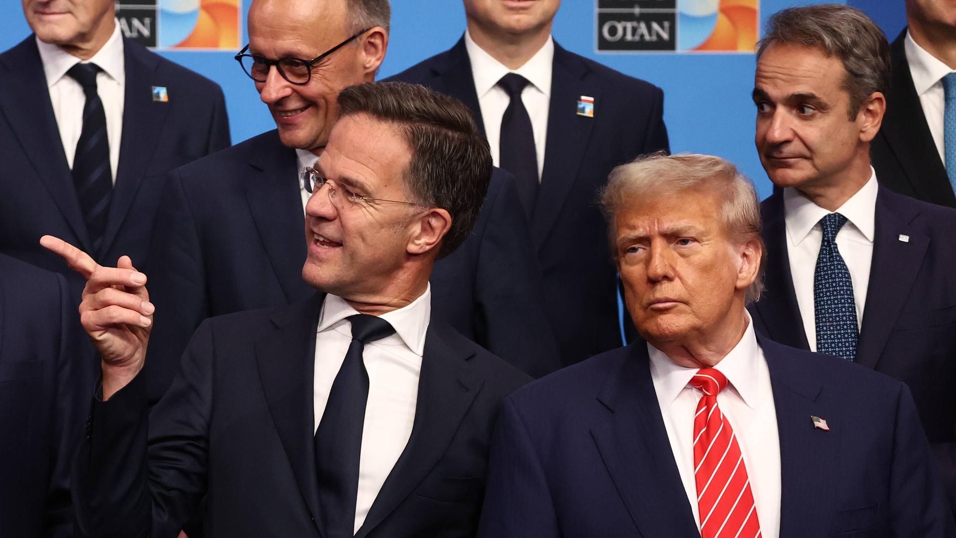 Beim NATO-Gipfel in Den Haag stehen Friedrich Merz, Mark Rutte und Donald Trump mit weiteren Politikern vor einer blauen Fotowand. Rutte zeigt mit dem Finger nach links.