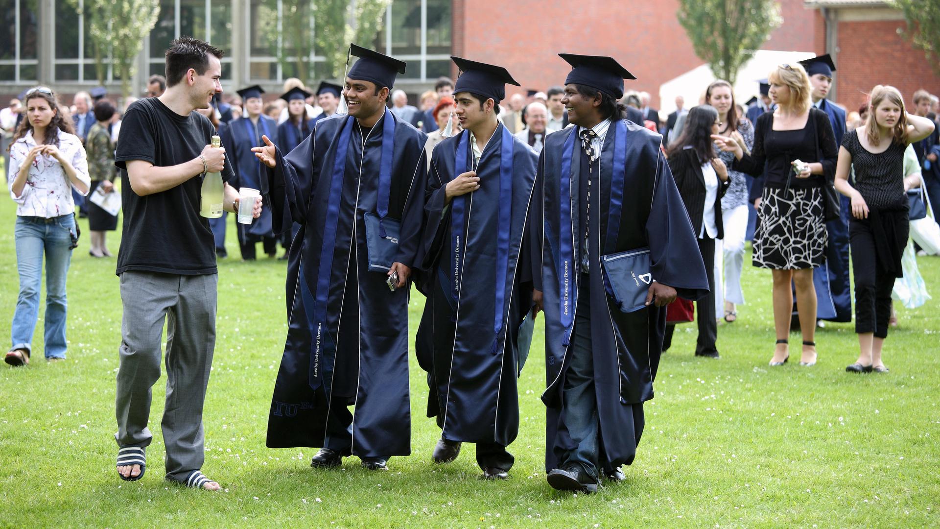 Abschlussfeier an der Uni in Bremen. Junge Menschen mit Doktorhüten gehen fröhlich über eine Wiese.