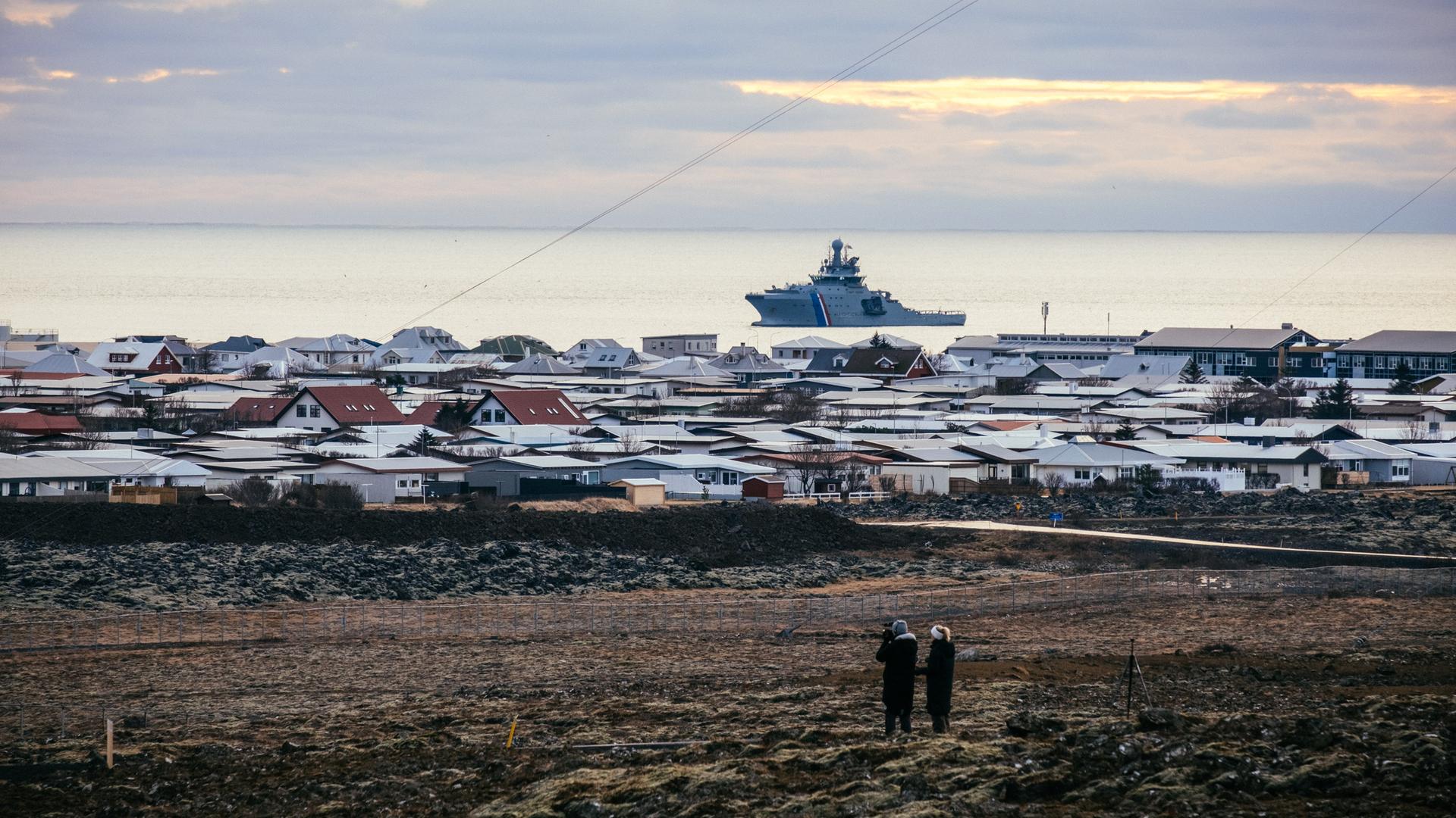 Ein Schiff der isländischen Küstenwache liegt vor Island im Meer. Auf dem Festland sind die Dächer von Häusern zu sehen.