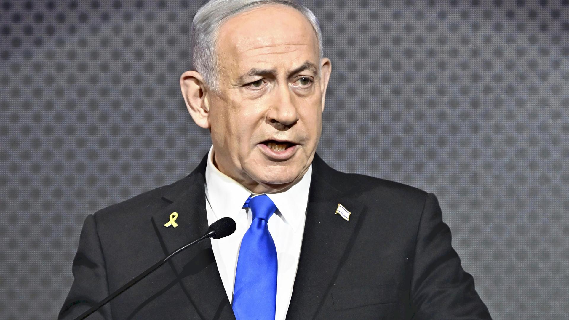 Der israelische Ministerpräsident Benjamin Netanjahu spricht im Anzug vor dunkelgrauem Hintergrund an einem Podium