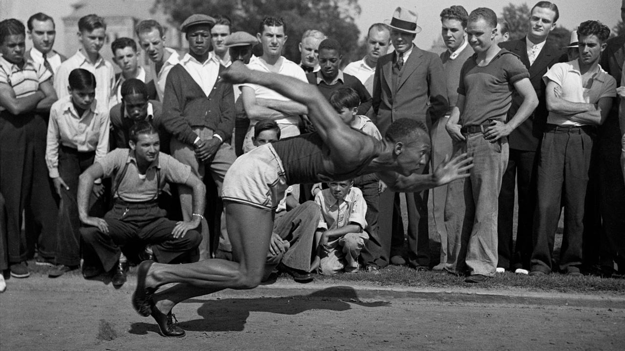 Ein Schwarz-Weiß-Foto zeigt Jesse Owens in einer dynamischen Pose beim Training. Eine Gruppe von Zuschauern beobachtet ihn dabei.