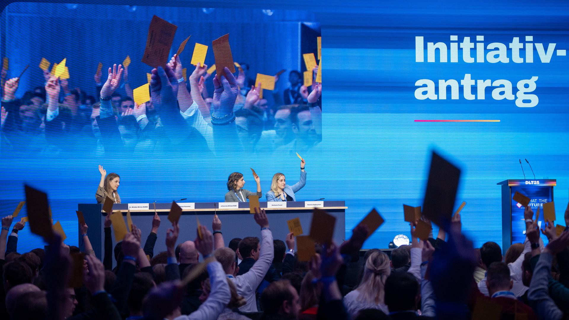 In einer Halle sitzen auf einem Podium auf dem Deutschlandtag der Jungen Union drei Personen, die die Hand gehoben haben oder Karten in die Höhe halten. Im Vordergrund sieht man Menschen von hinten im Publikum, die gelbe Karten in die Höhe halten.