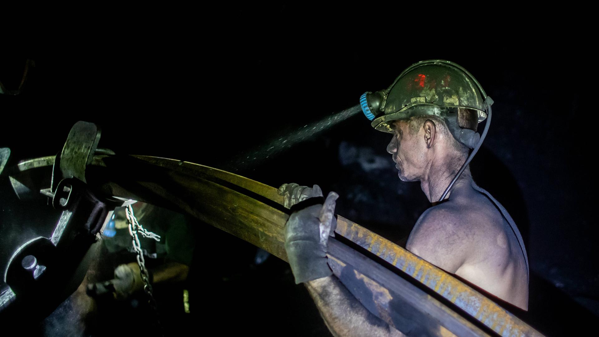 Ein mit Kohlenstaub bedeckter Bergmann trägt mit freiem Oberkörper ein schweres Metallrohr. Von seinem gelben Helm strahlt die Grubenlampe in die Dunkelheit der Kohlenmine. Stonava near Karvinâa, Czech Republicâ, January 22, 2025. 