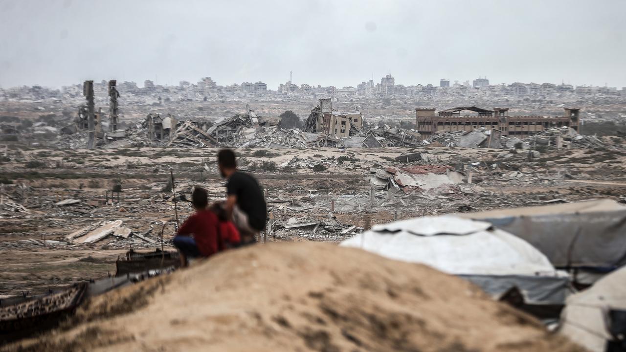 Zwei Personen, die von hinten zu sehen sind, darunter ein Kind, sitzen auf einem Hügel und schauen auf die zerstörte Stadt von Gaza