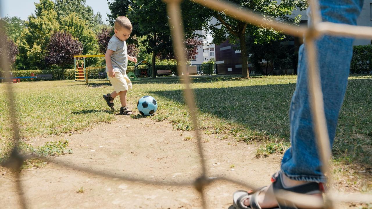 Ein kleiner Junge spielt Fußball, während ein Erwachsener im Tor steht. Von ihm sieht man nur ein Bein im Bild.