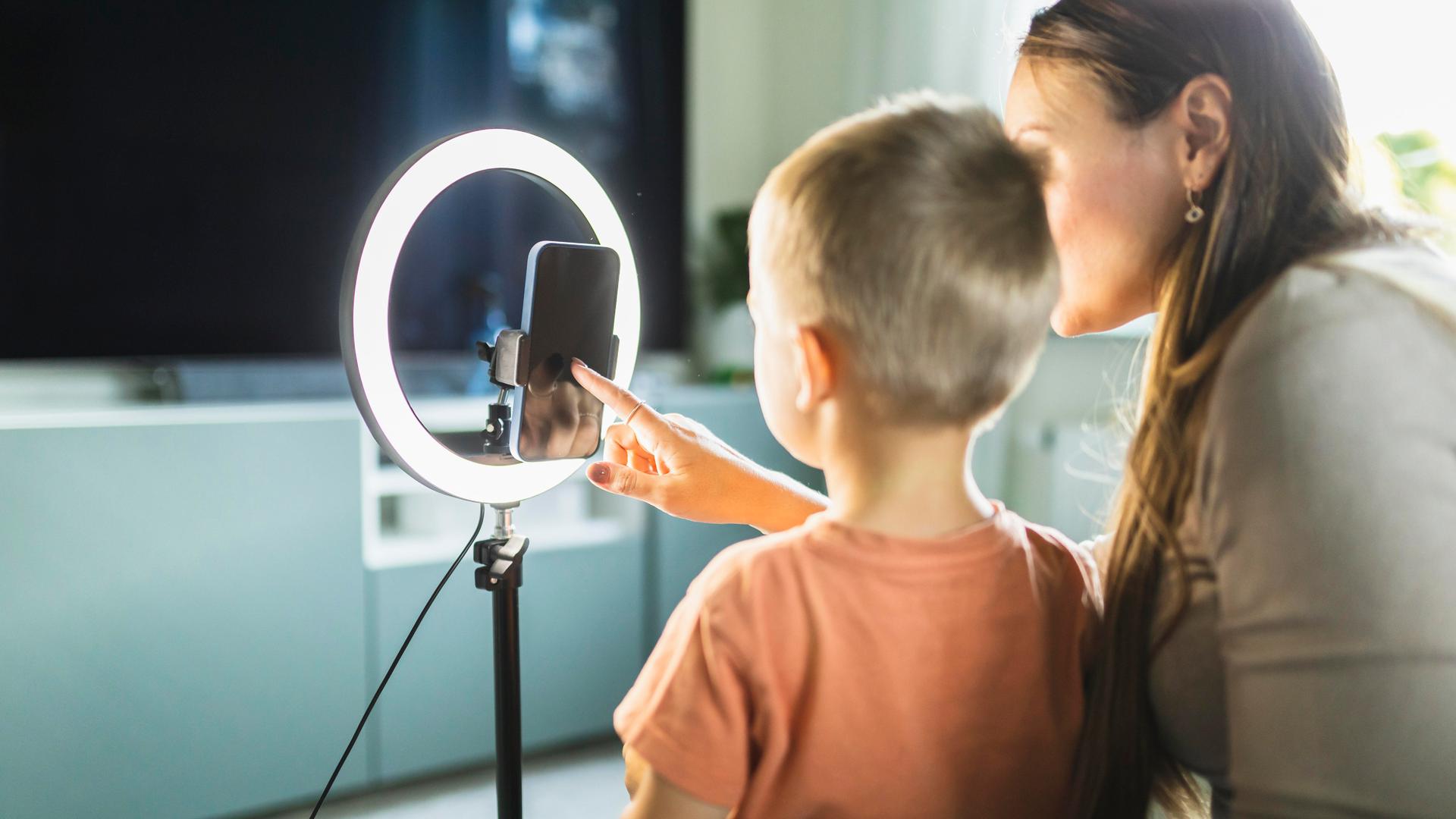 Ein Kind sitzt vor einem mit Stativ und Ringlicht ausgestatteten Smartphone, das von einer erwachsenen Frau neben ihm bedient wird.
