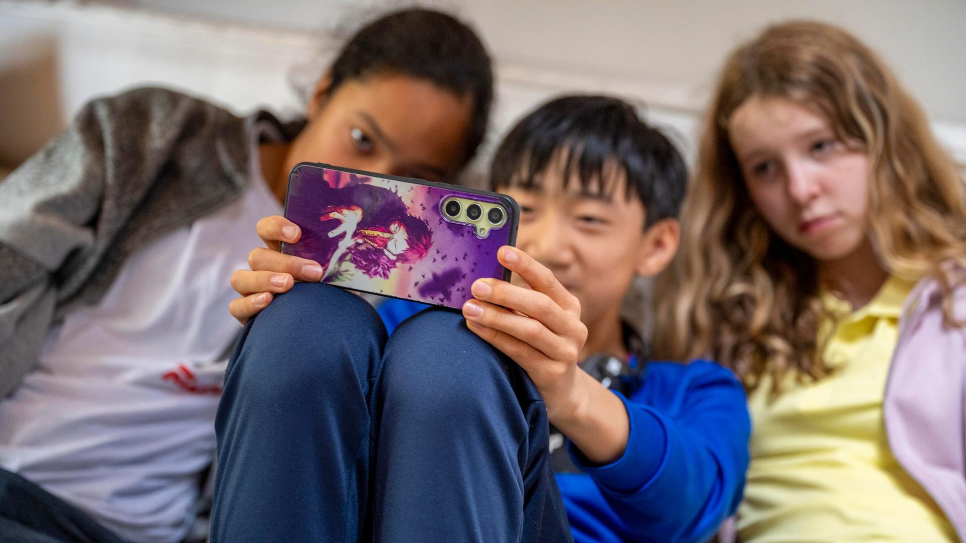 Zwei Mädchen und ein Junge sitzen dicht nebeneinander auf der Couch und machen mit einem Smartphone ein Selfie von sich