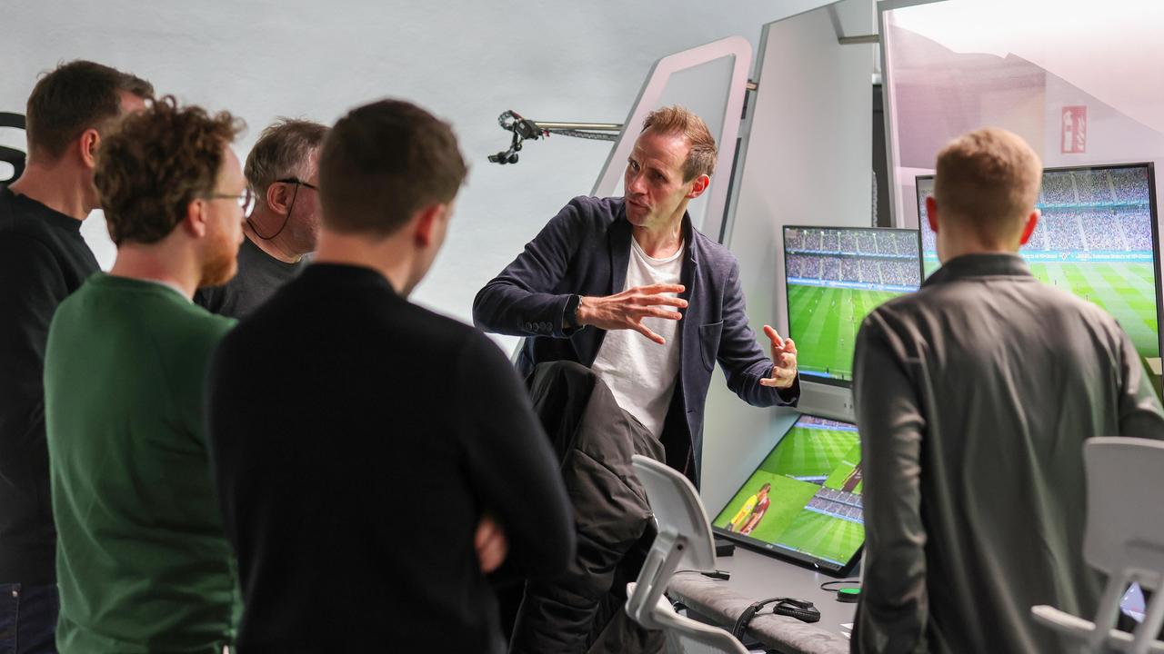 Eine Personengruppe steht dem Betrachter mit dem Rücken zugewandt vor einer Wand mit mehreren Monitoren, auf denen ein Fußballfeld im Stadion zu sehen ist. Eine Person erklärt gestikulierend das Prozedere.