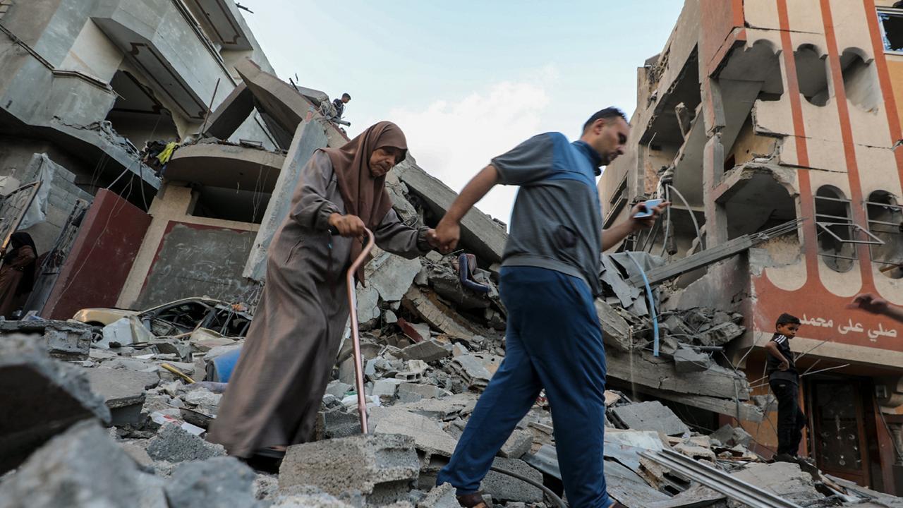Ein Mann führt eine ältere Frau am Gehstock aus dem Schutt eines zerstörten Gebäudes heraus.