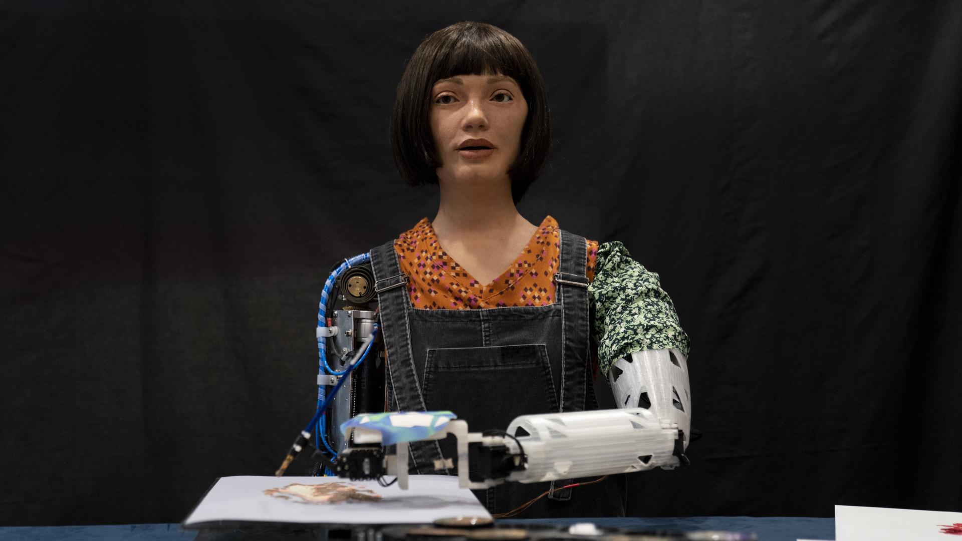 Roboter-Künstlerin Ai-Da malt Bilder mithilfe von KI in der British Library in London. Wir sehen einen Roboter mit weiblich wirkendem Gesicht. Vor ihr liegt ein Bild, auf dem sie mit dem linken Roboterarm malt. 