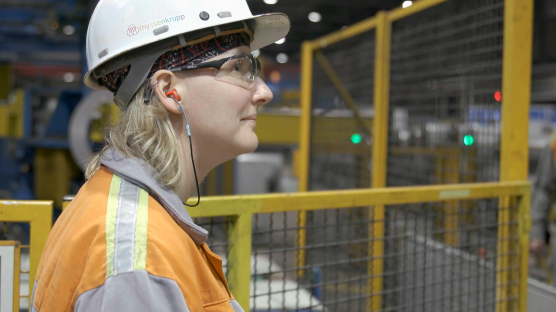 Ann Catherine Meyer steht mit Schutzhelm und Arbeitsjacke in einer Produktionshalle von Thyssenkrupp und blickt nach rechts 
