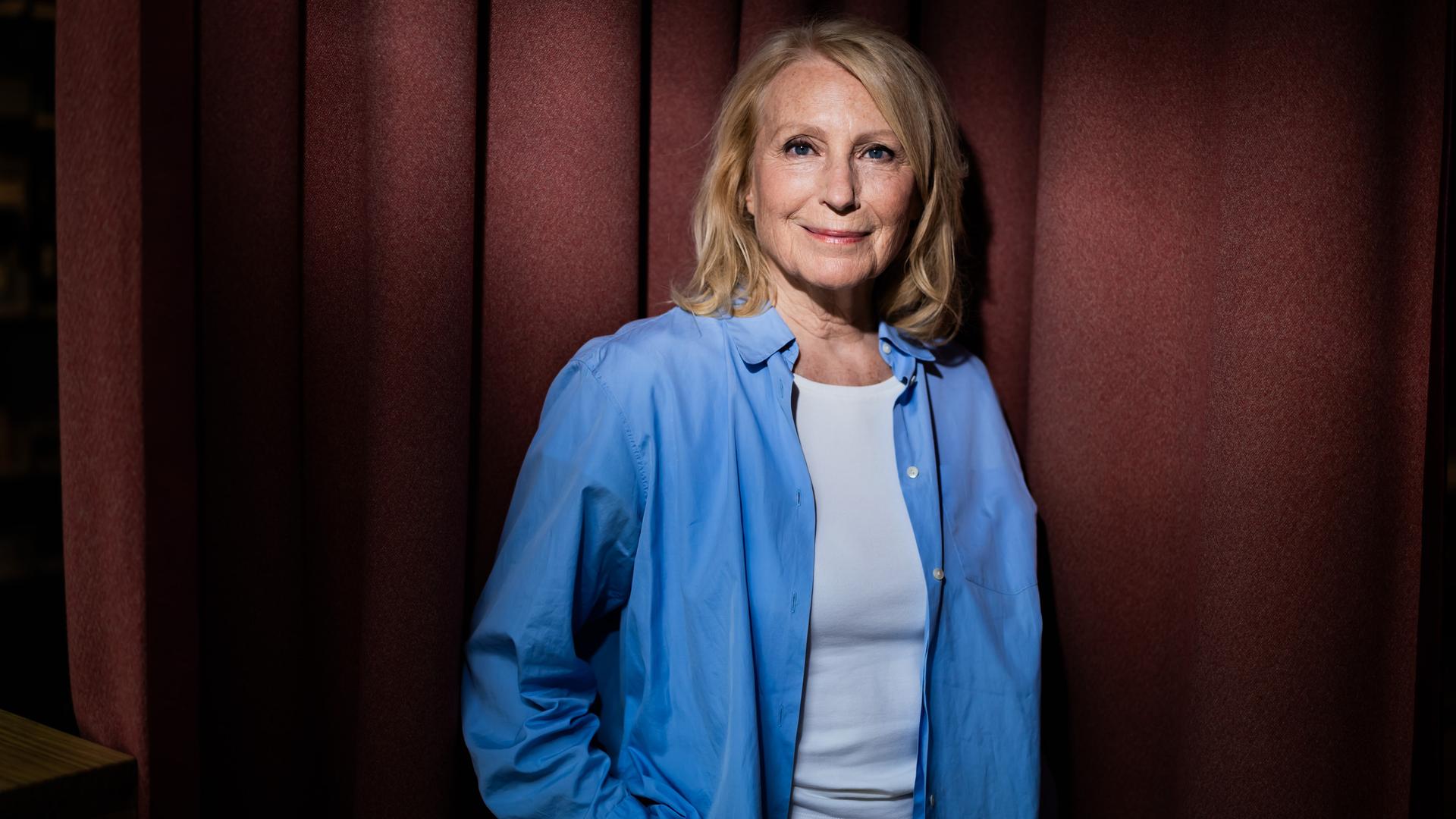 Maren Kroymann, Schauspielerin, steht während eines Fototermins in weißem T-Shirt und offenem blauen Hemd vor einem Vorhang und lächelt in die Kamera.