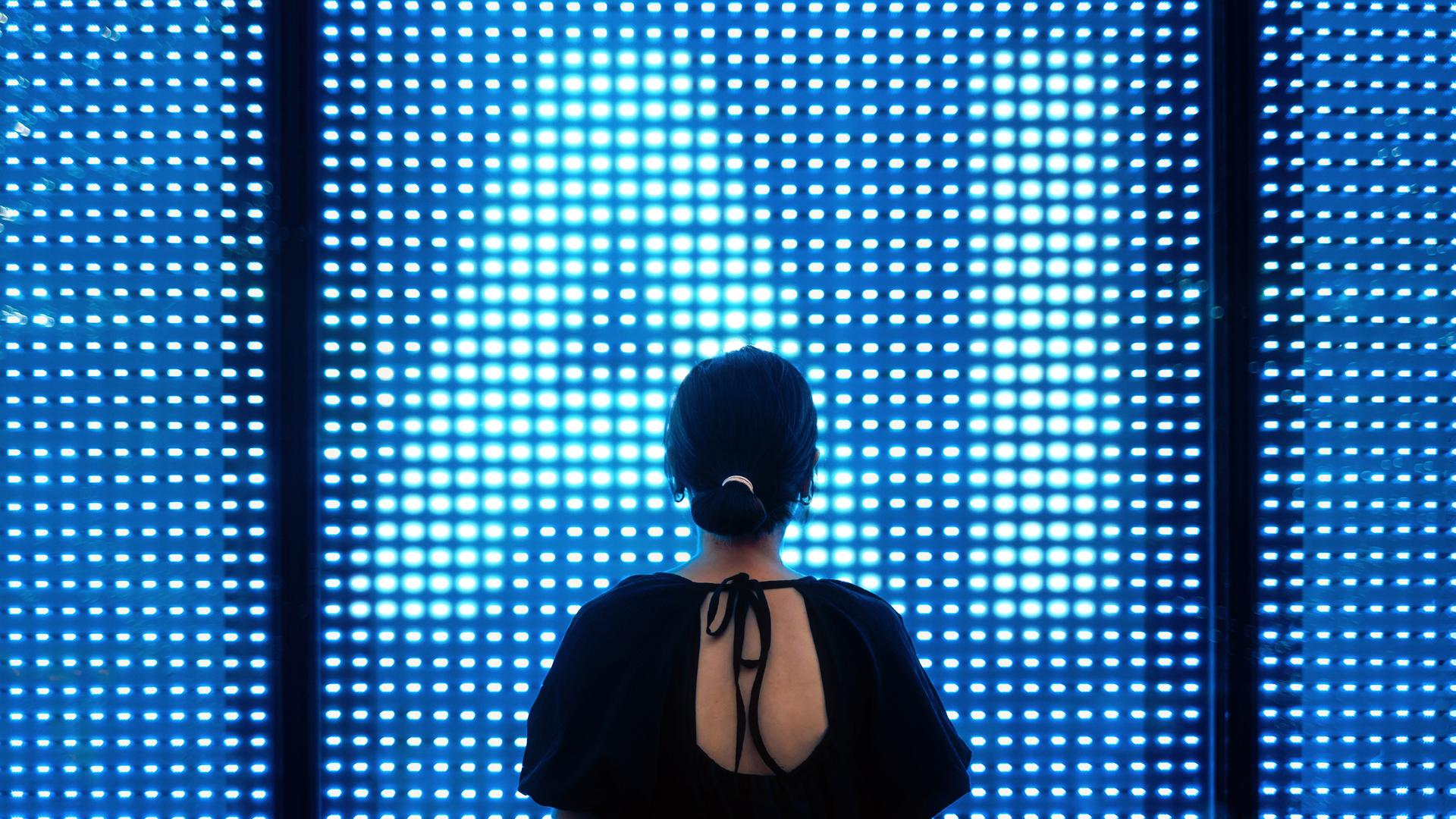 Eine Frau steht dem Betrachter mit dem Rücken zugewandt vor einer erleuchtenden LED Wand, deren Lichtpunkte die Buchstaben "AI" formen.